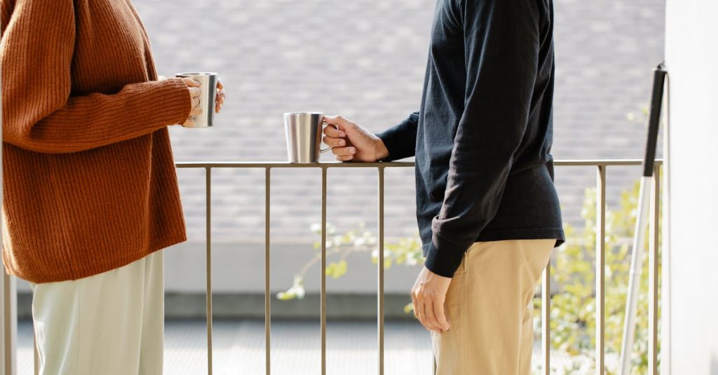 Two people on a balcony holding mugs, enjoying a casual conversation in soft sunlight.